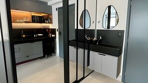 A modern kitchen with black countertops, a built-in oven, and a large mirror above the sink. - Get By Side (João Pessoa)
