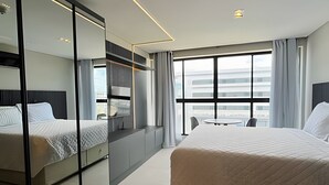 A modern hotel room with a large bed, a desk, a chair, a mirror, and a large window with a view. - Get By Side (João Pessoa)
