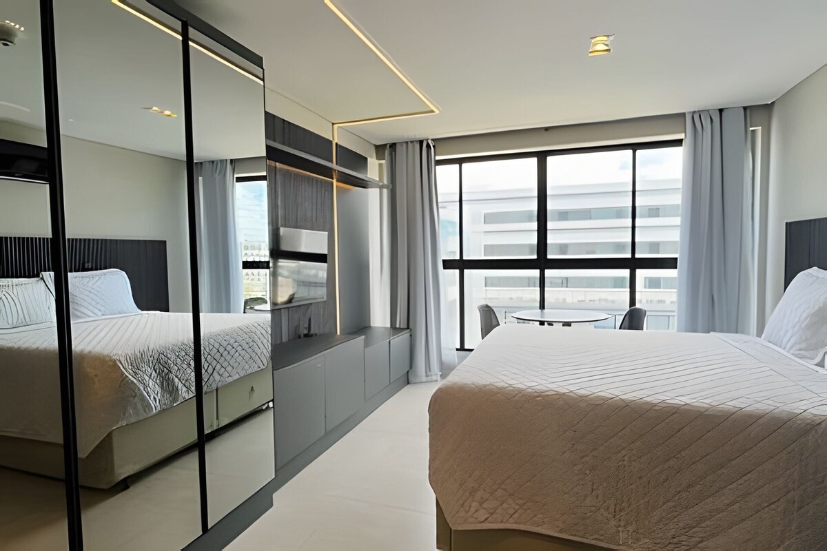 A modern hotel room with a large bed, a desk, a chair, a mirror, and a large window with a view.