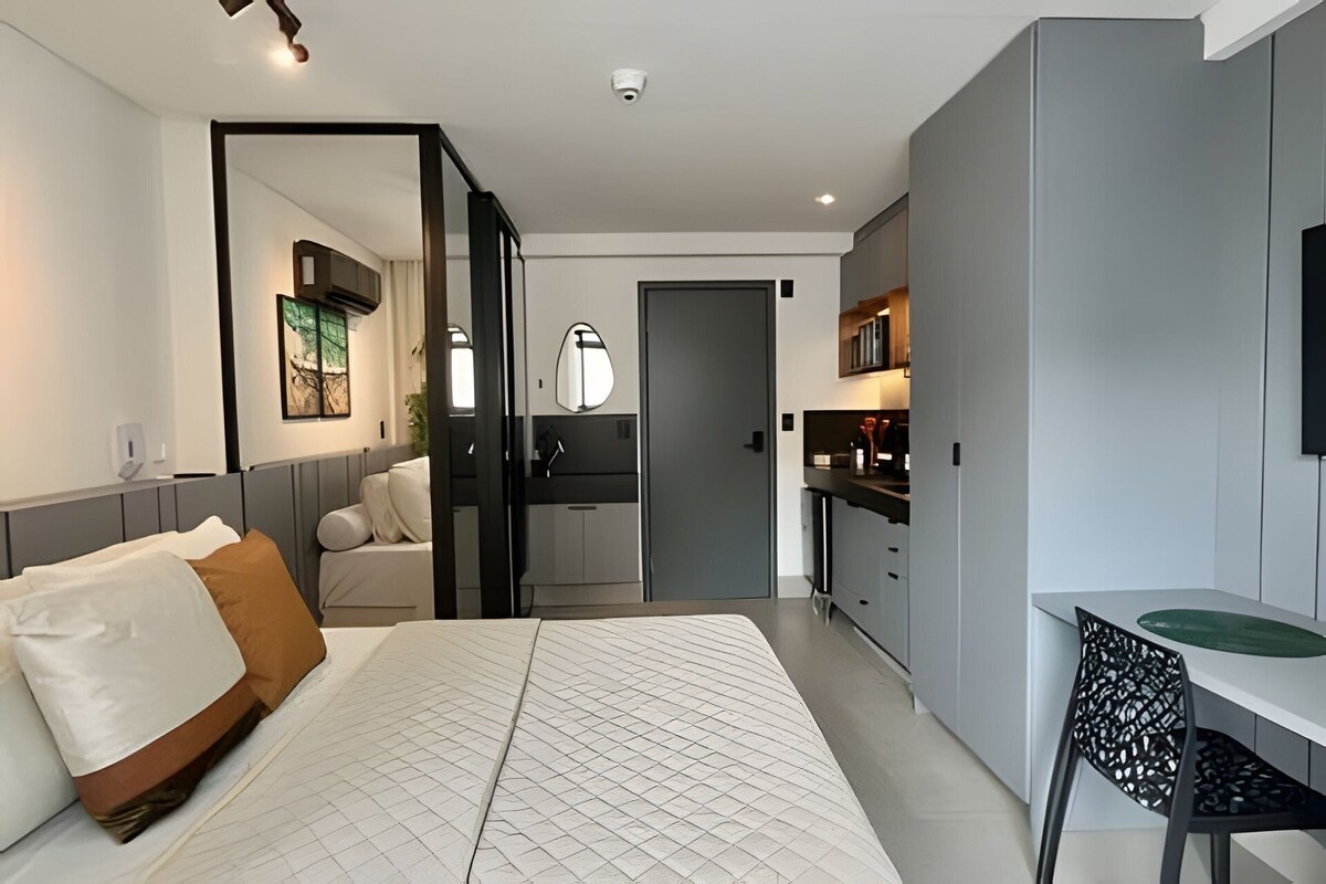 A modern hotel room with a bed, a desk, a chair, a kitchenette, and a bathroom.