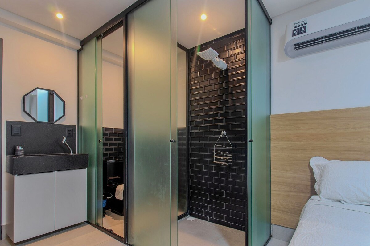 A modern bedroom with a black brick wall, a wooden headboard, a white bed, a bathroom with a black sink and mirror, and a glass door closet.
