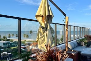 Uma varanda com vista para o mar, um guarda-sol amarelo e um espaço de estar com almofadas.