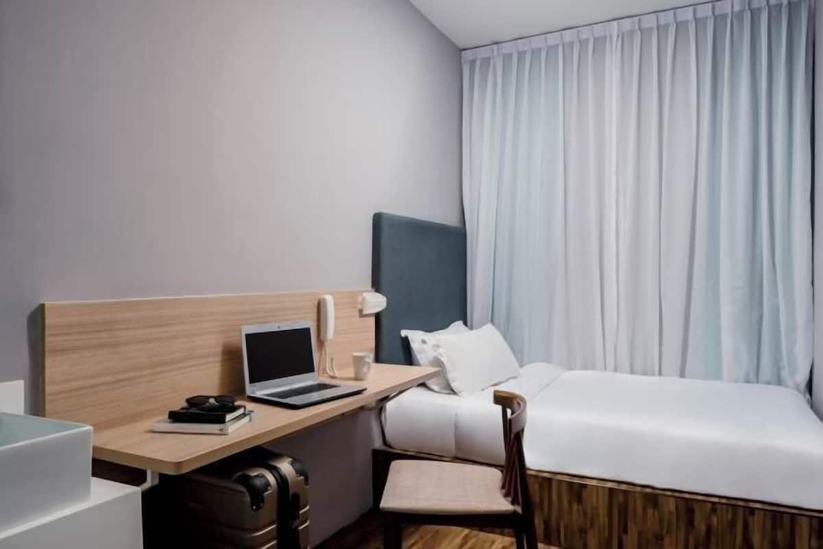 A hotel room with a bed, a desk with a laptop, a chair, a suitcase, and a window with curtains.