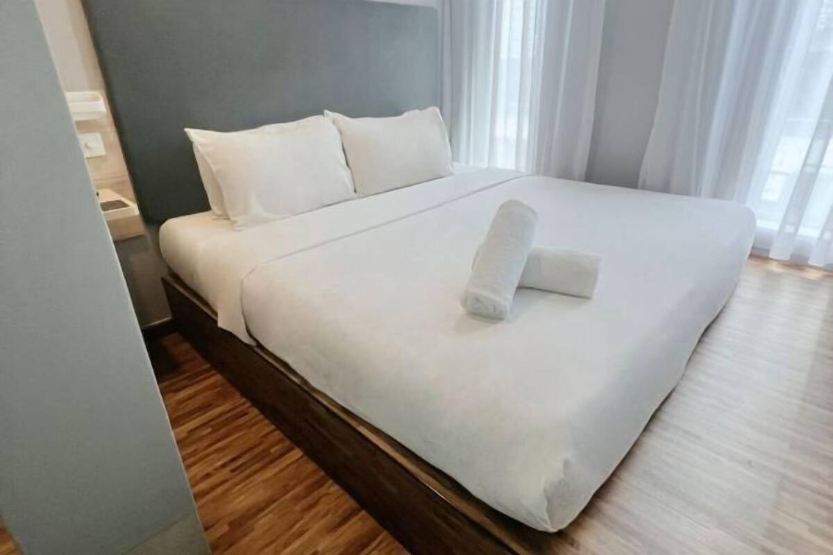 A neatly made bed with white linens and a rolled towel, a wooden headboard, and a light-colored wall.
