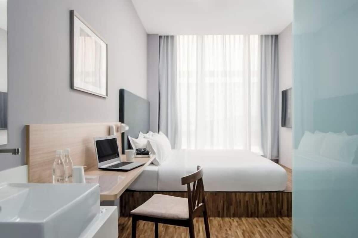 A hotel room with a bed, a desk with a laptop, a chair, a bedside table with a lamp, a window with curtains, and a wall-mounted TV.