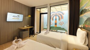 Hotelkamer met twee bedden, een televisie aan de muur, een bureau en uitzicht op een palmboom door het raam.