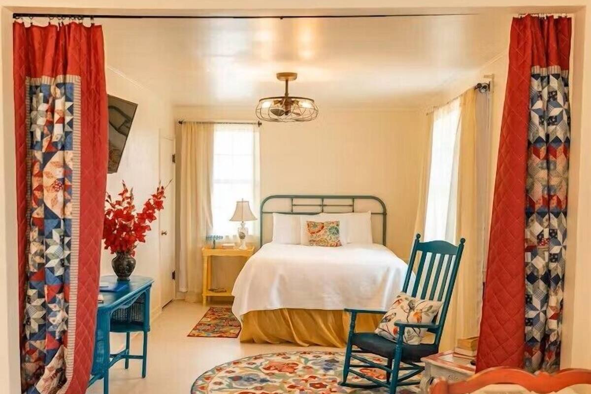 A bedroom with a bed, a chair, a table, a lamp, and a window.