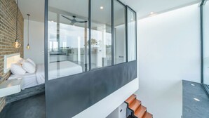 Un dormitorio moderno con pared de vidrio, cama con ropa blanca, pared de ladrillo y ventilador de techo.