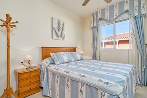 A bedroom with a wooden headboard, a bed with a floral-patterned bedspread, a nightstand with a lamp, and a window with curtains.