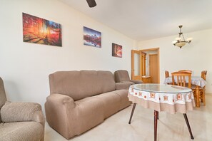 A living room with a sofa, a glass table, and two chairs. There are paintings on the wall and a chandelier.