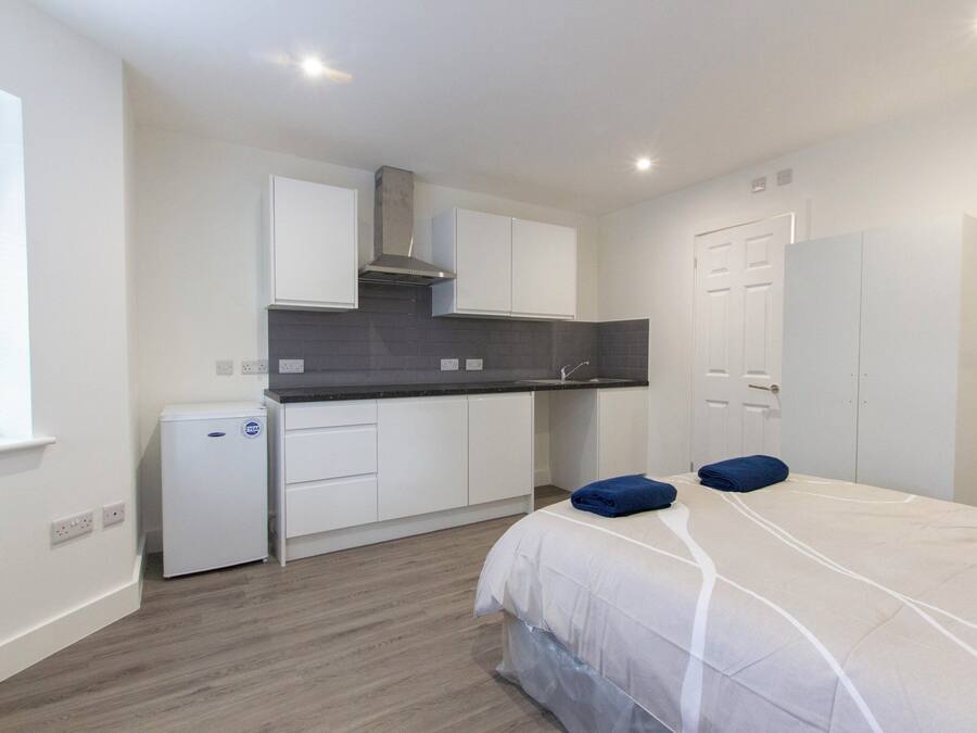 A compact hotel room with a kitchenette, a bed with a white duvet, and a small refrigerator.