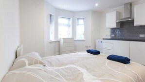 Una habitación de hotel moderna y compacta con cama, una kitchenette pequeña y dos ventanas que aportan luz natural.