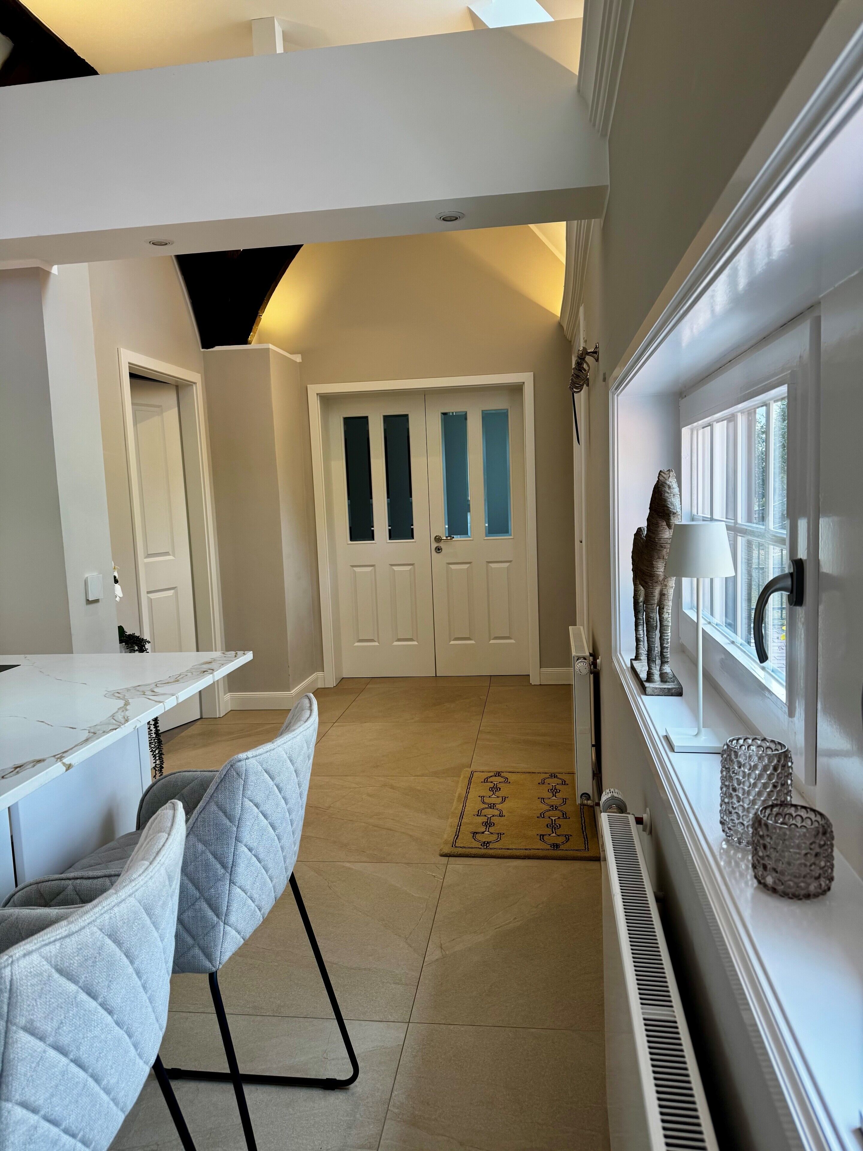 A modern kitchen with a marble countertop, two upholstered chairs, a white door with frosted glass panels, and a window with decorative elements.