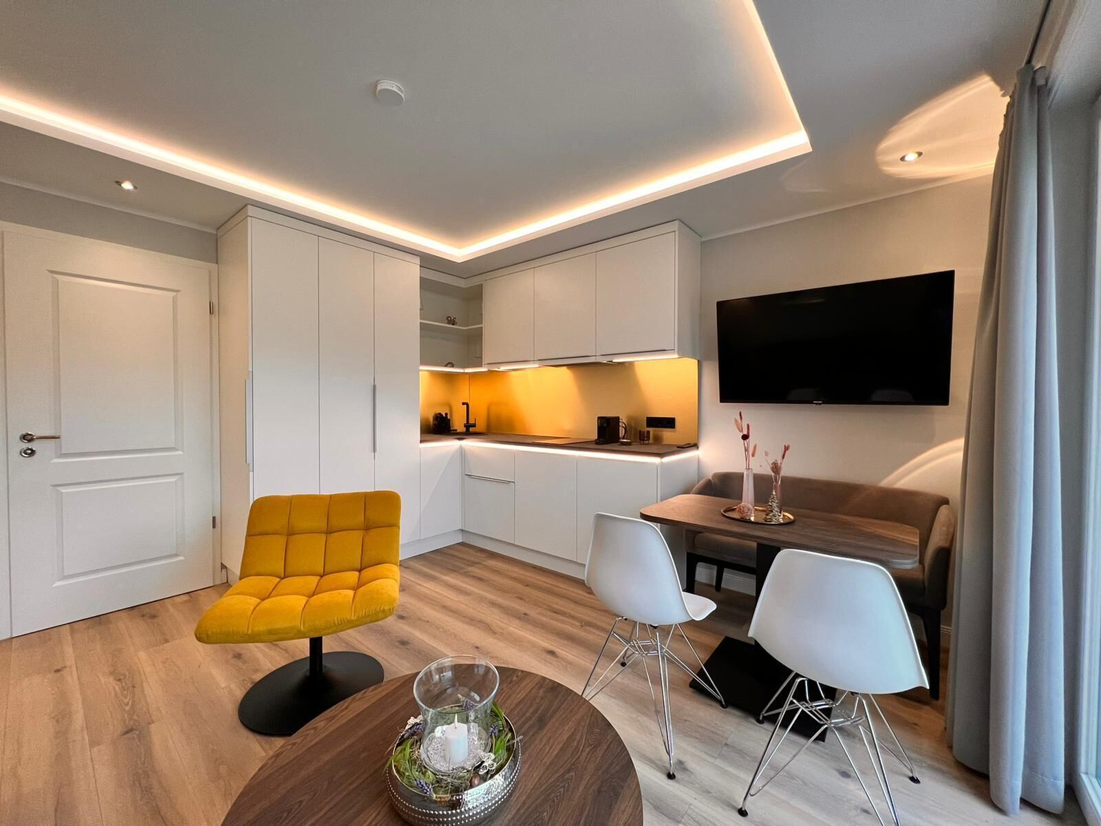 A modern living room with a yellow armchair, a dining area with white chairs, a wooden coffee table, and a kitchen area with white cabinets and a TV mounted on the wall.