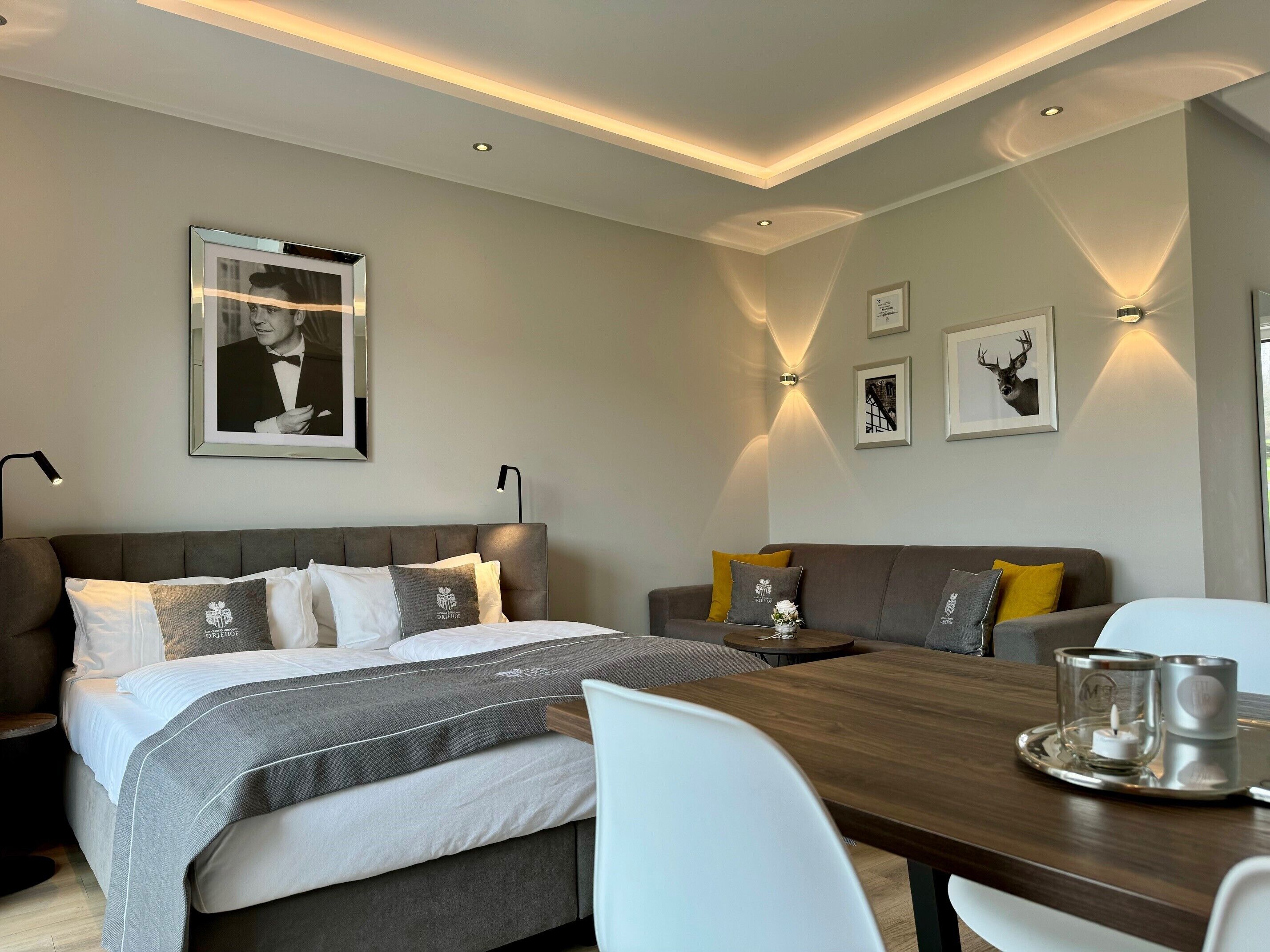 A modern hotel room with a bed, sofa, dining table, and wall-mounted artwork.