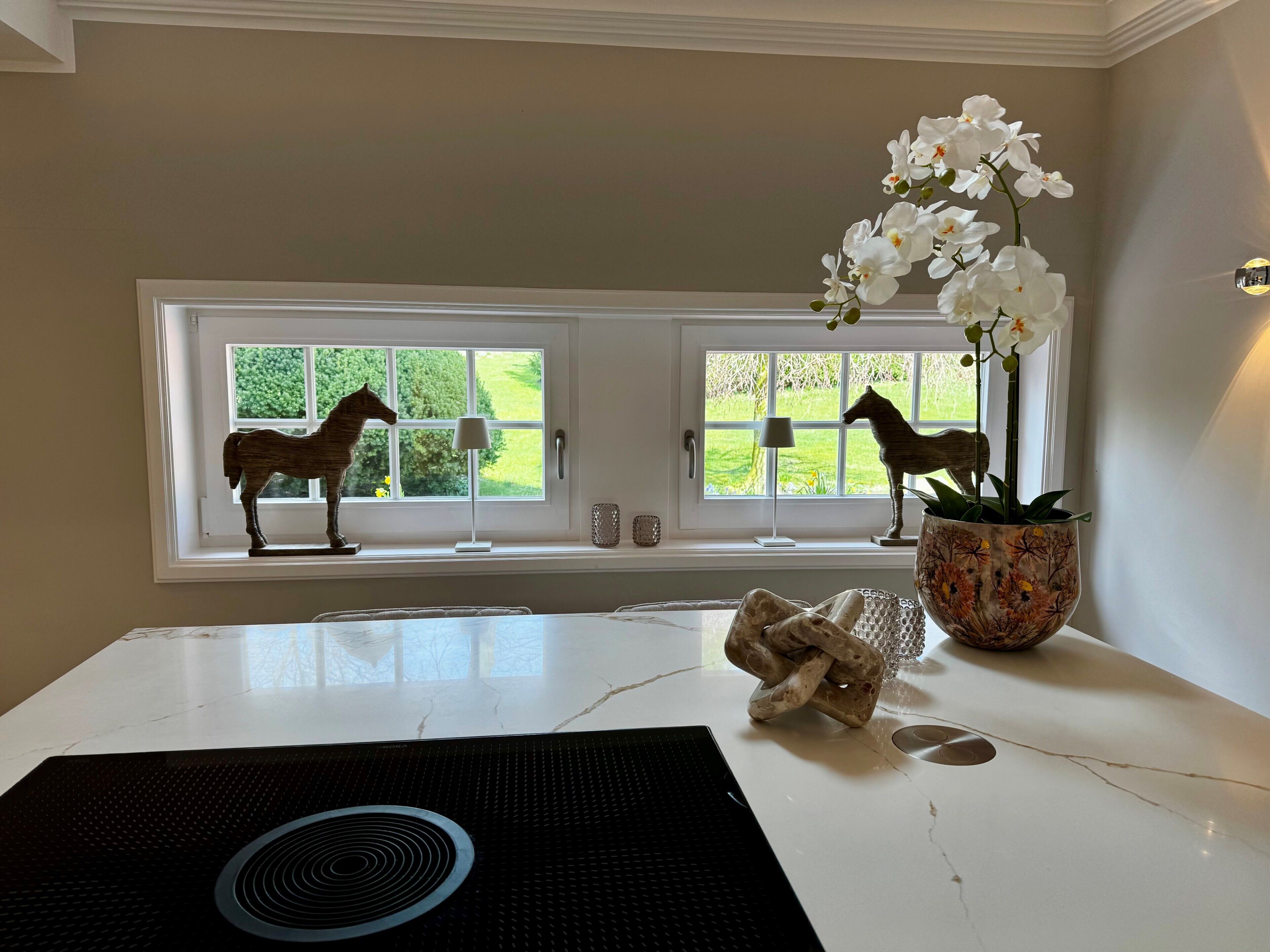 Una cocina con encimera de mármol, placa de inducción negra, dos esculturas con forma de caballo en los alféizares de las ventanas y una orquídea en maceta.