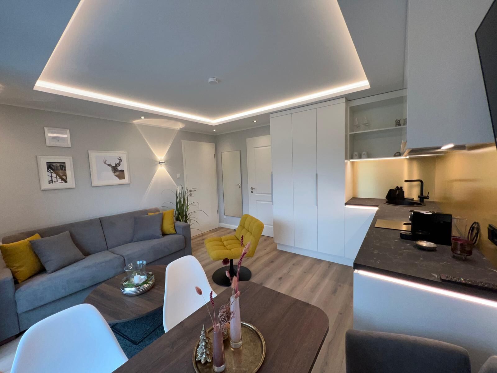 Un salon moderne avec un canapé, un fauteuil jaune, une table à manger agrémentée d’un vase, et un coin cuisine avec des placards et une cuisinière.