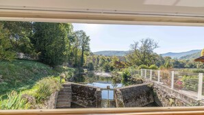 Une vue à travers une fenêtre sur un paysage naturel, avec un étang, des marches en pierre et une clôture blanche.