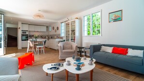 A modern living room with a grey sofa, a red and white pillow, a dining area with a table and chairs, and a kitchen in the background.