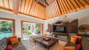 A spacious living room with a large sofa, a coffee table, and a television set. There is a pool visible through the glass doors. - The Complete Guide to Renting Your Exclusive Holiday Villa in Kabupaten Badung with Private Pool and Fully Staffed (Bali)