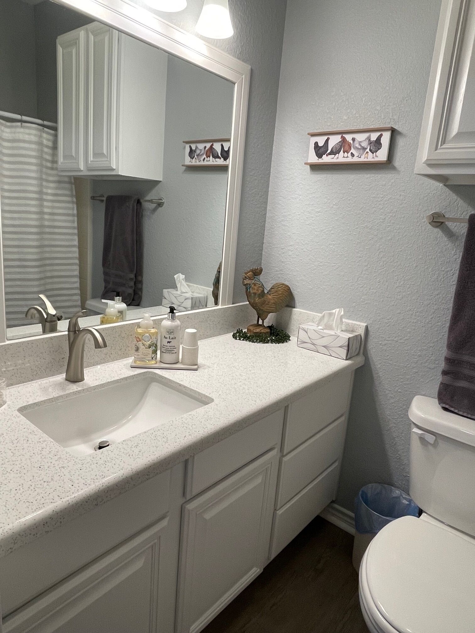 Une salle de bain avec un lavabo blanc, un miroir et des toilettes. On y trouve des articles de toilette sur le lavabo, un porte-serviettes et un tableau encadré au mur.