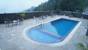 Una piscina rectangular con borde alicatado, rodeada de baldosas de piedra y una pared de piedra. Varias sillas y mesas con plantas en macetas se encuentran alrededor de la piscina.