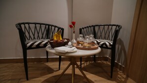 Una mesa de comedor con una cesta de frutas, un jarrón con flores y una bandeja con frutos secos y semillas. Dos sillas con cojines estampados están dispuestas alrededor de la mesa.