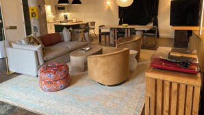 A modern living room with a sofa, ottoman, and coffee table, a dining area with a table and chairs, and a kitchen in the background.