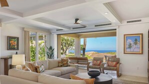 A spacious living room with a large sofa, armchairs, and a coffee table. There is a view of the ocean through large windows. - Wai'ula'ula J101 Three Bedroom (Kamuela)