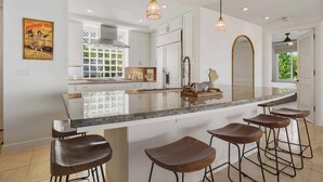 A modern kitchen with a large island, bar stools, pendant lights, and a decorative mirror. - Wai'ula'ula J101 Three Bedroom (Kamuela)