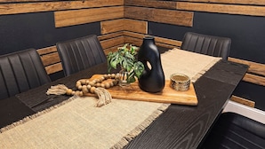 Uma mesa de jantar com acabamento em madeira escura, cercada por cadeiras de couro preto. Um jogo americano trançado e uma pequena planta em vaso preto estão sobre a mesa.