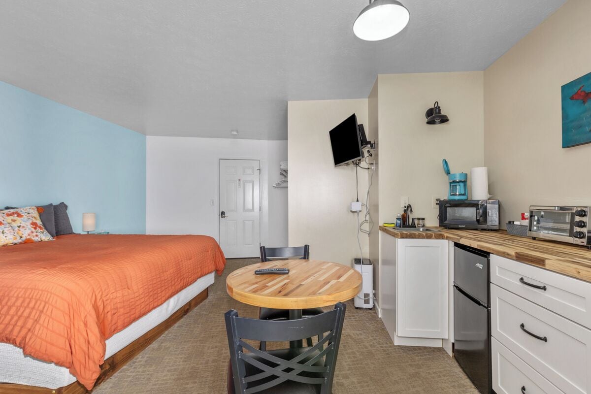 A compact hotel room with a bed, a small round table, a kitchenette, and a TV mounted on the wall.