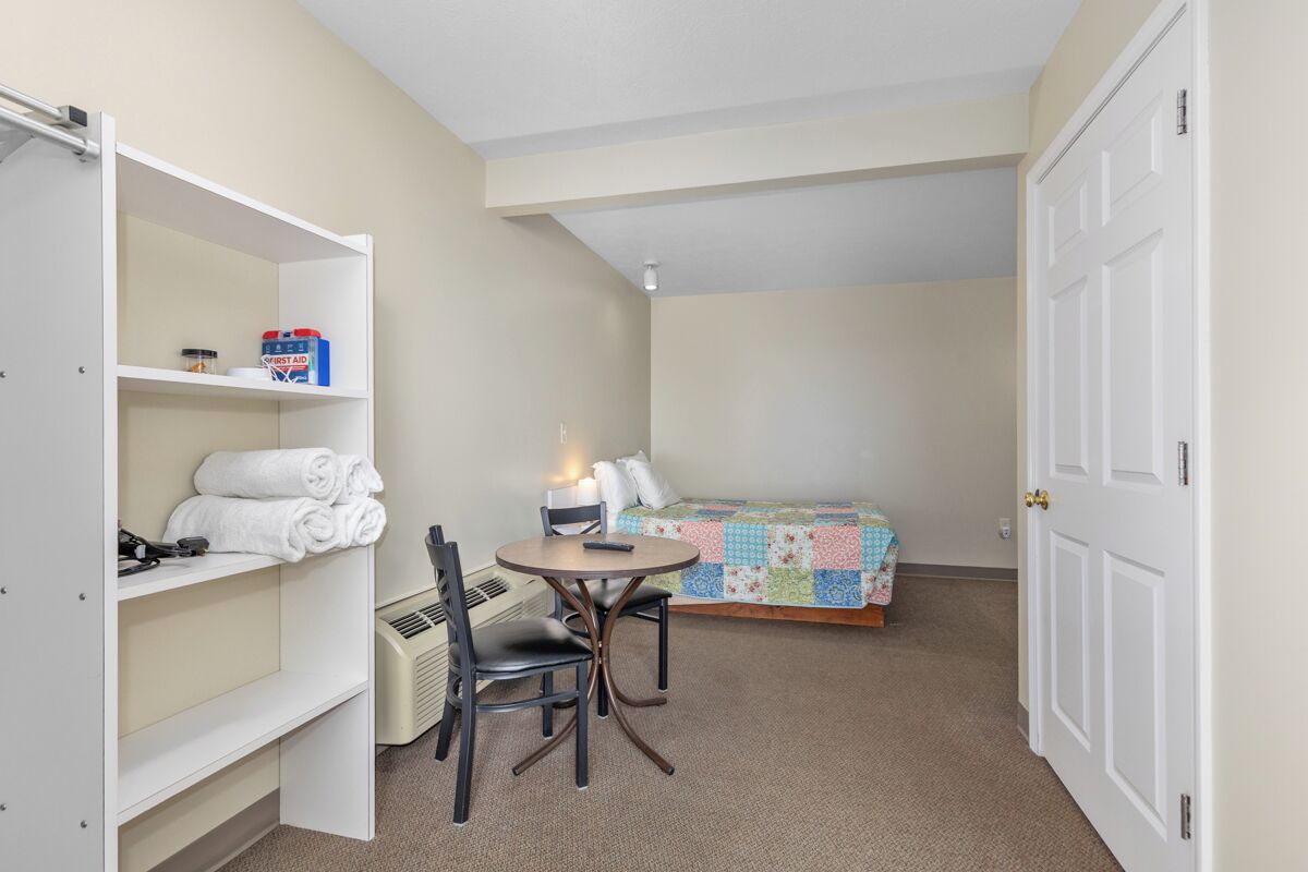 Een kleine kamer met een bed, een tafeltje met stoelen, een verwarming en een kledingkast.