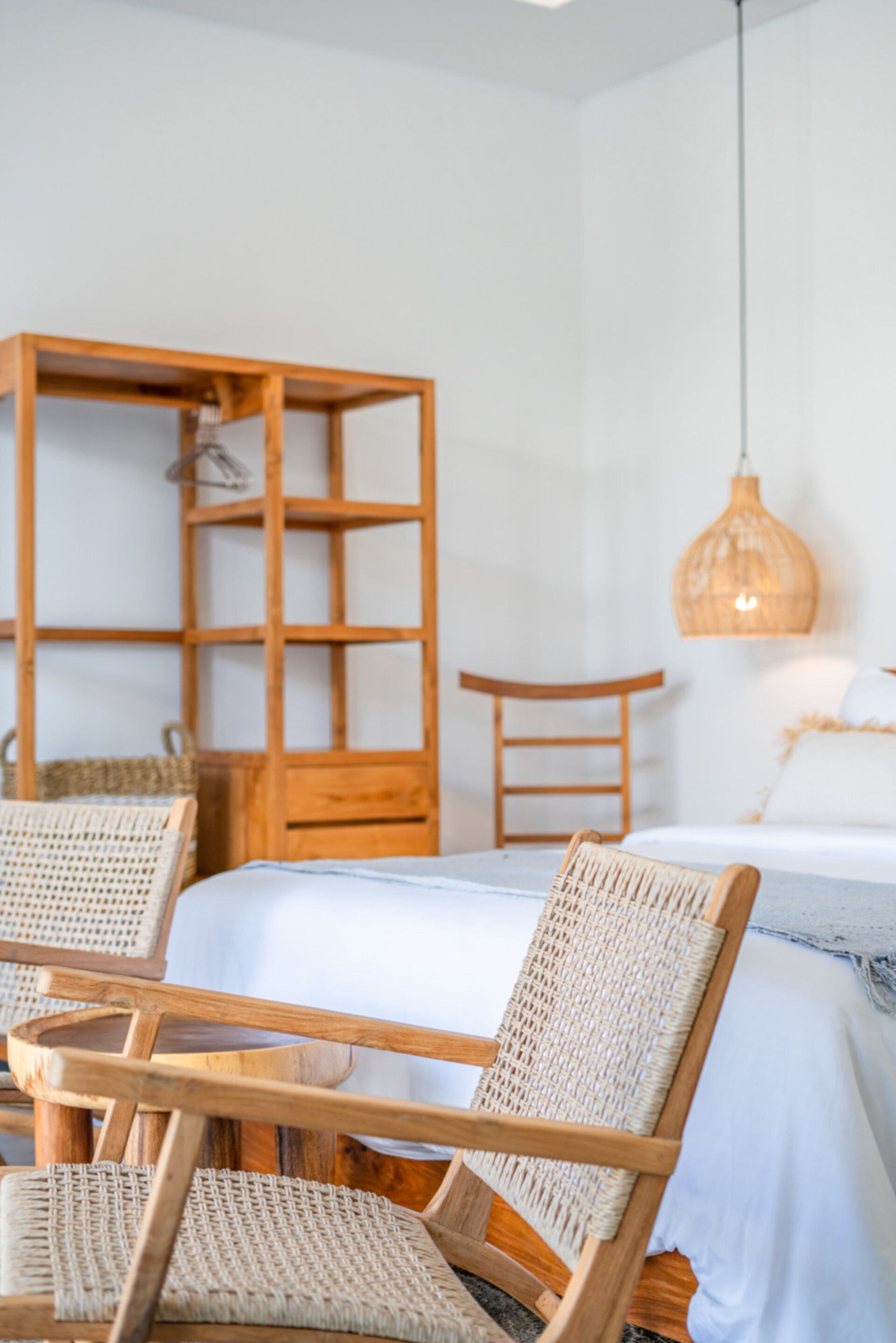 Una habitación con una cama, dos sillones de mimbre, un aparador de madera y una lámpara colgante.