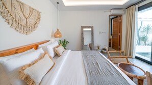 A modern hotel room with a large bed, a wooden headboard, a macrame wall hanging, a hanging pendant light, a large mirror, a wooden chair, and a view of the outdoors through a large window.