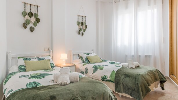 Une chambre avec deux lits, chacun recouvert d’une couvre-lit à motifs de feuilles vertes, une table de chevet avec une lampe et une décoration en macramé accrochée au mur.