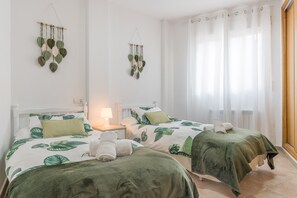 Ein Zimmer mit zwei Betten, beide mit einem Bettbezug mit grünem Blattmuster, einem Nachttisch mit Lampe und einer Wanddekoration aus Makramee.
