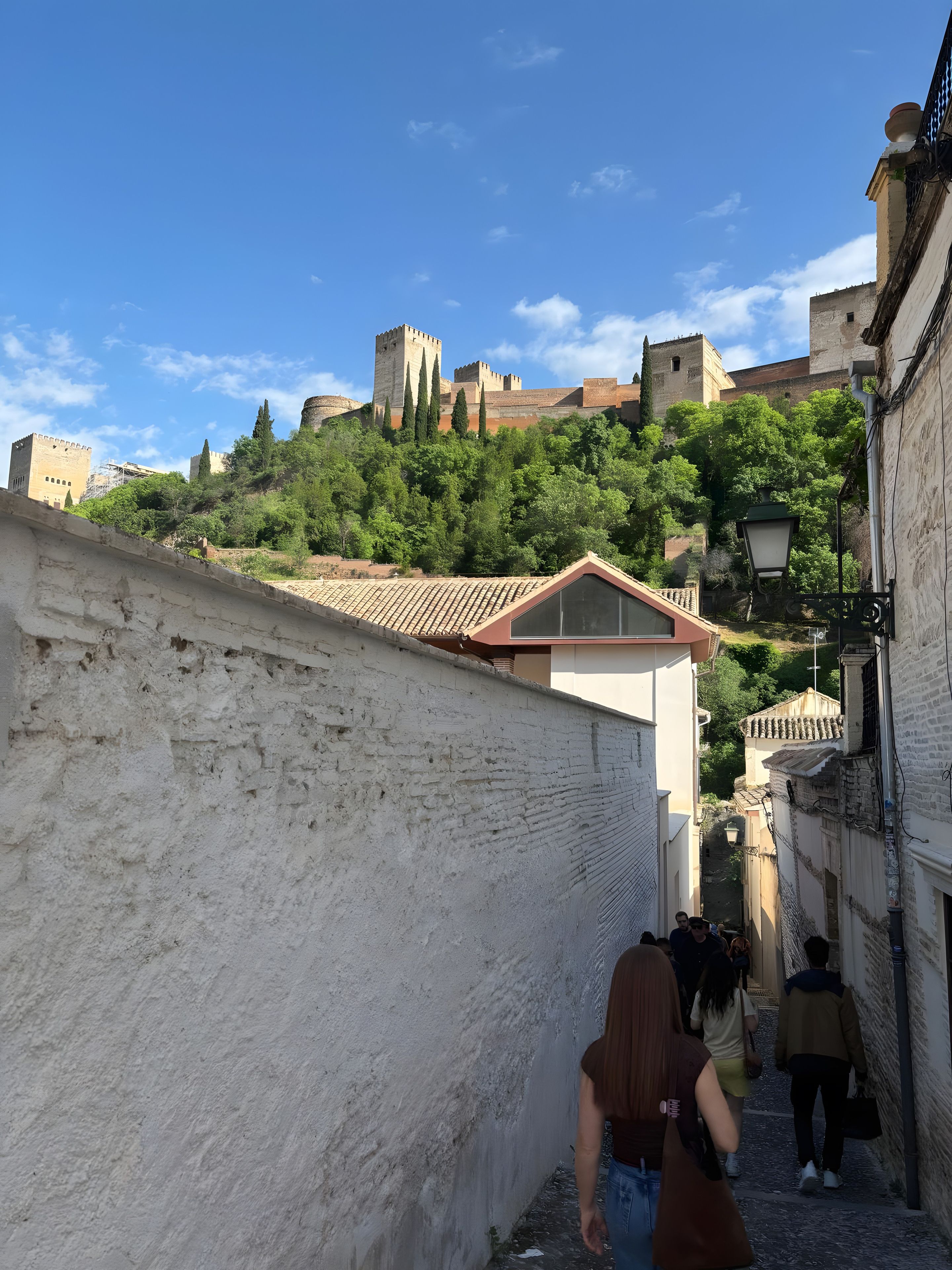 A narrow street with a historical building on a hill, a white wall, and a few pedestrians.