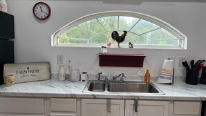 A kitchen with a marble countertop, a sink, a window with a rooster decoration, and a clock on the wall.