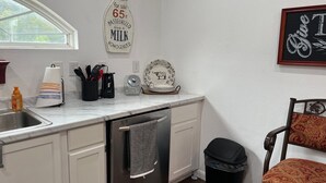 A kitchen with a stainless steel dishwasher, a window, a milk sign, and a chair with a patterned cushion.
