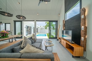 Uma sala de estar moderna com um sofá cinzento, uma mesa de centro e um rack para TV. Uma piscina é visível através das portas de vidro.