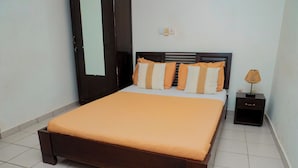 Una cama bien hecha con cabecera marrón y ropa de cama naranja, una mesita de noche pequeña con una lámpara, y un enchufe de pared.