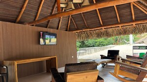 A covered outdoor seating area with wooden furniture, a television screen, and a view of the beach. - hacienda hotel texas (Durania)