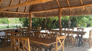 A covered outdoor seating area with wooden tables and chairs, a thatched roof, and a view of greenery and a sandy beach. - hacienda hotel texas (Durania)