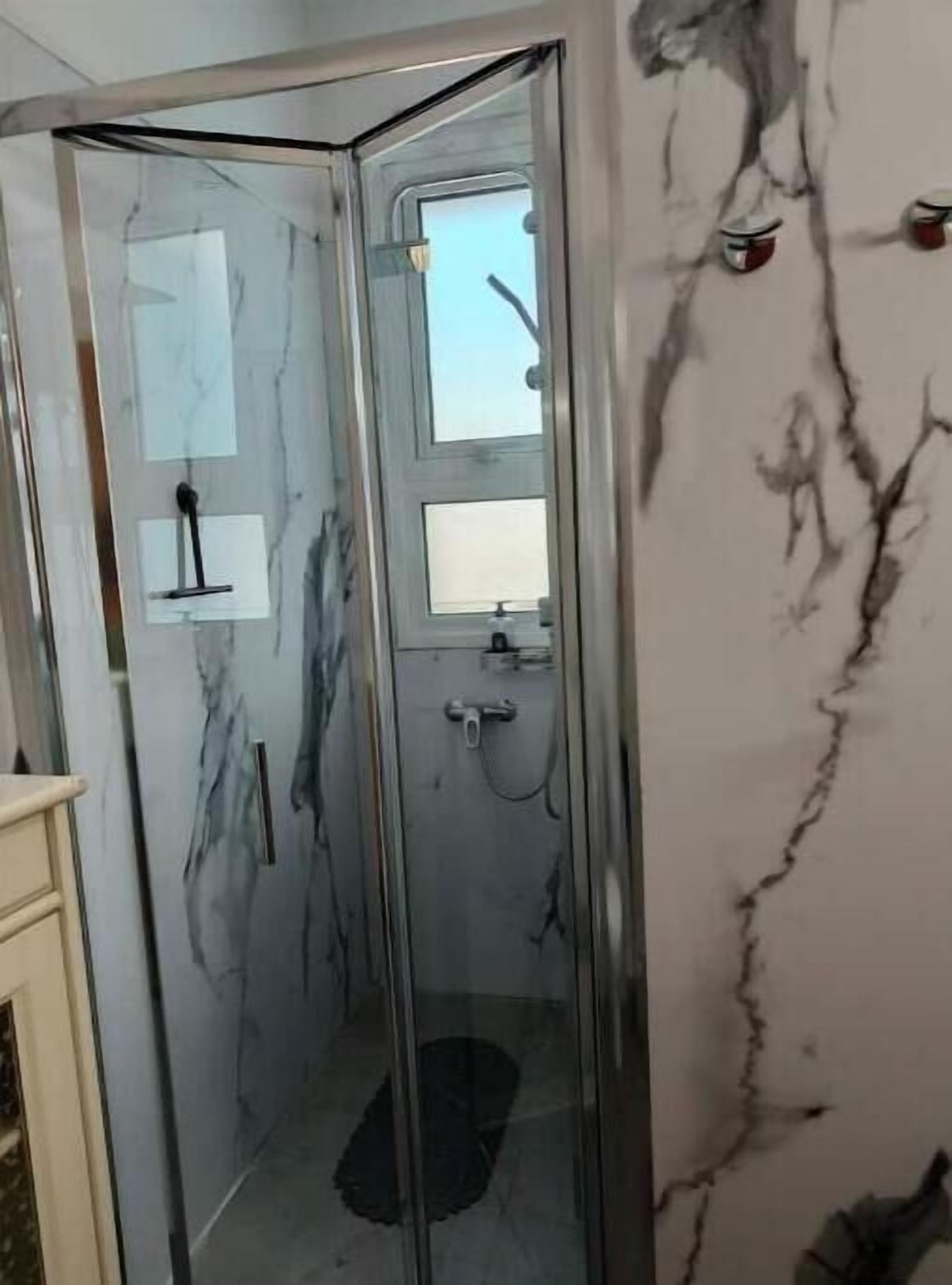A bathroom with a glass shower enclosure, marble walls, and a small shelf with toiletries.