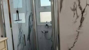 A bathroom with a glass shower enclosure, marble walls, and a small shelf with toiletries.