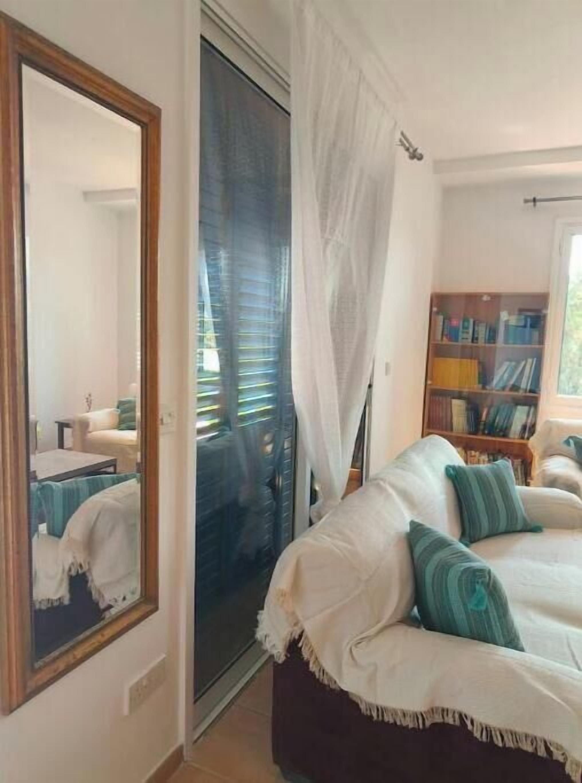 A bedroom with a large mirror, a sliding glass door leading to a balcony, a bed with white bedding and green pillows, and a bookshelf filled with books.