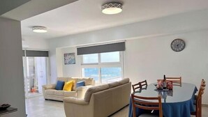 Ein geräumiger Wohnbereich mit einer Couch, einem Esstisch und Stühlen. Ein Fenster bietet einen Ausblick, an der Wand hängt eine Uhr und es gibt eine Deckenleuchte.