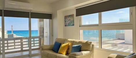 A spacious living room with a beige sofa, a smaller couch, and a large window offering a view of the sea.