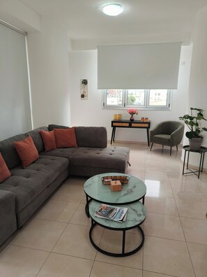 A living room with a sectional sofa, two round tables, a chair, a small side table, and a window with a view.